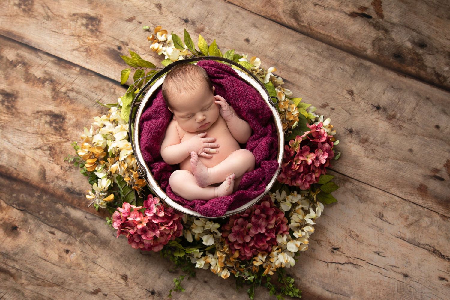 Creative newborn photography with baby nestled among flowers in purple basket on rustic wood floor.