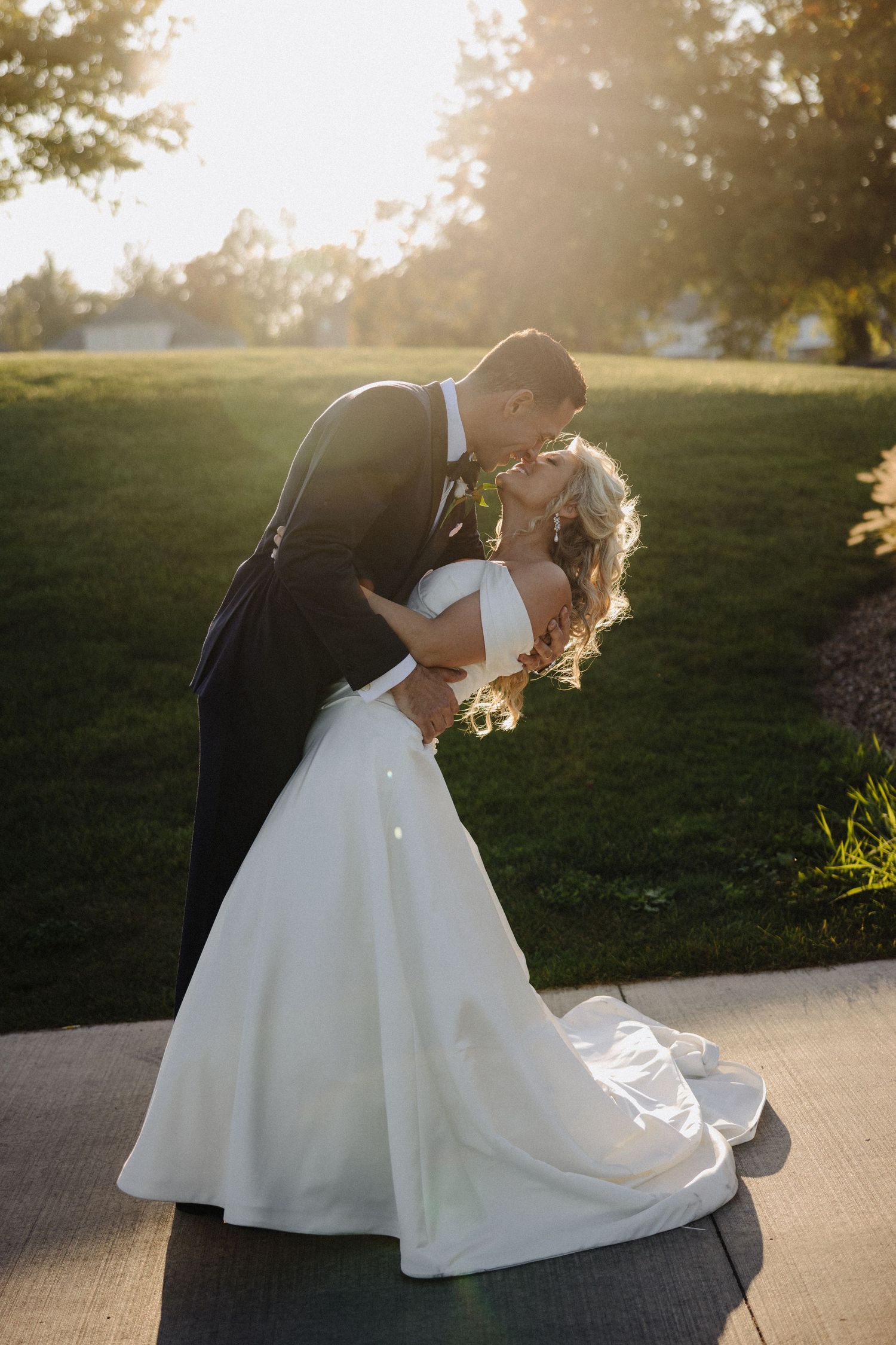 Romantic wedding photo of a couple in formal attire dipping and kissing at sunset on a garden path.