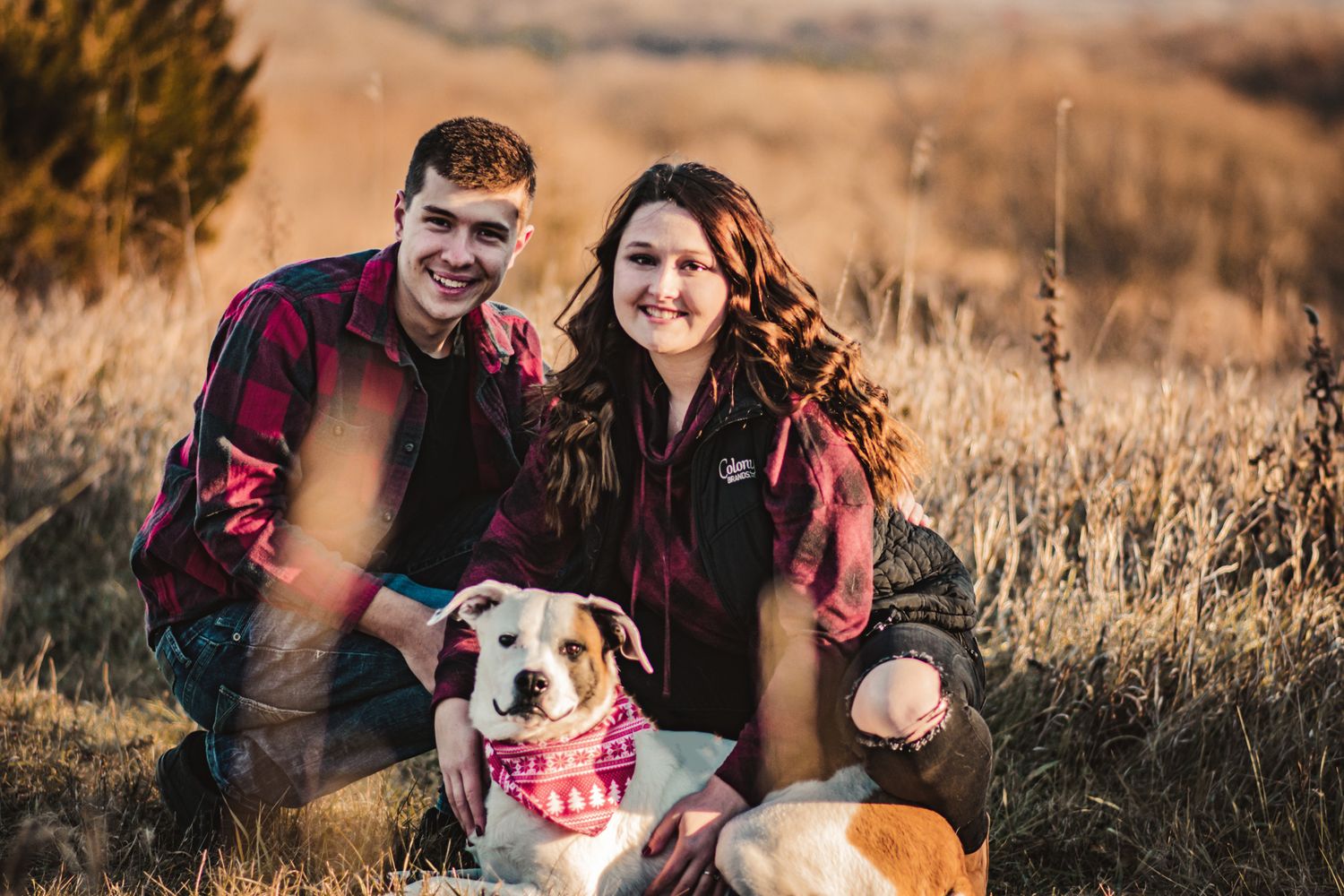 Happy couple posing with their smiling dog in a field wearing matching red plaid shirts.