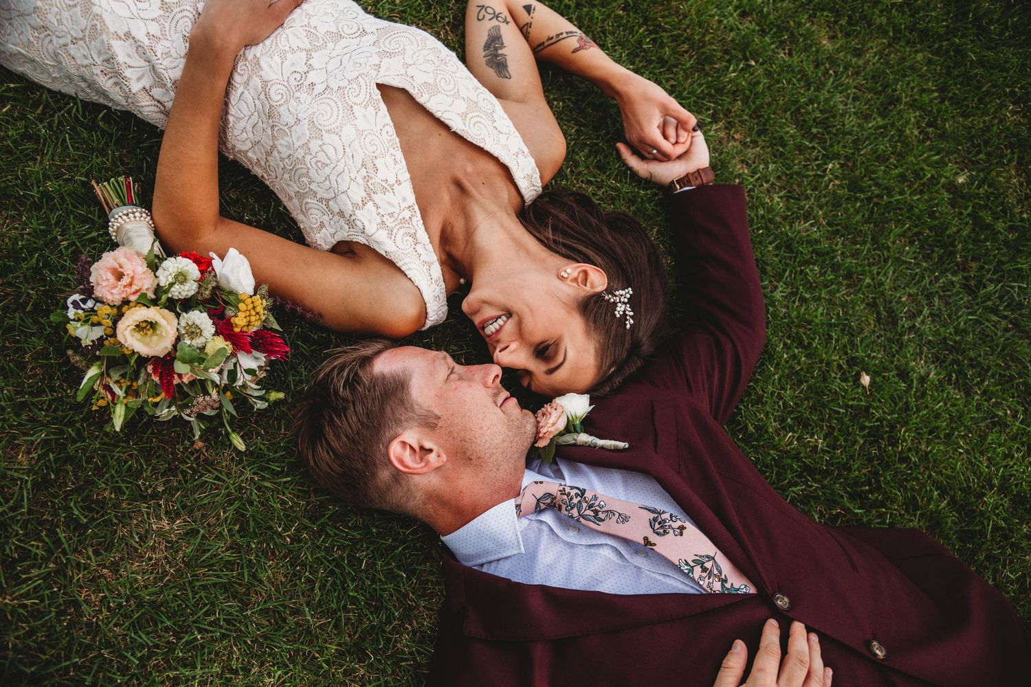 Overhead view of couple in wedding attire lying on grass with floral bouquet.