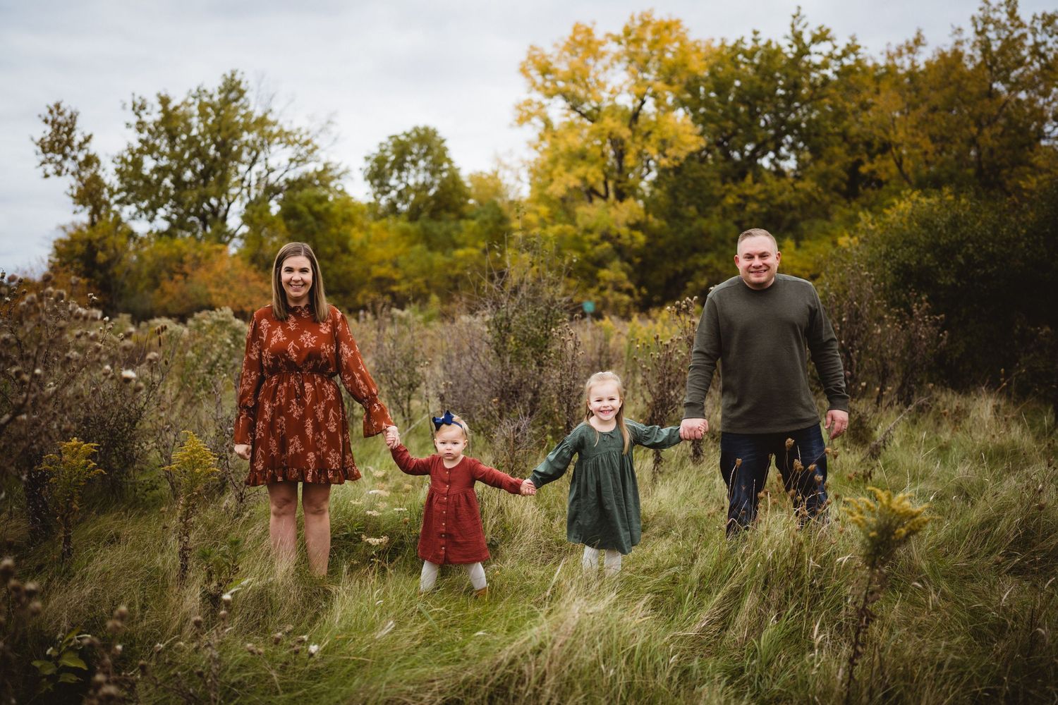 Family standing together in tall grass with autumn trees and yellow foliage in the background.