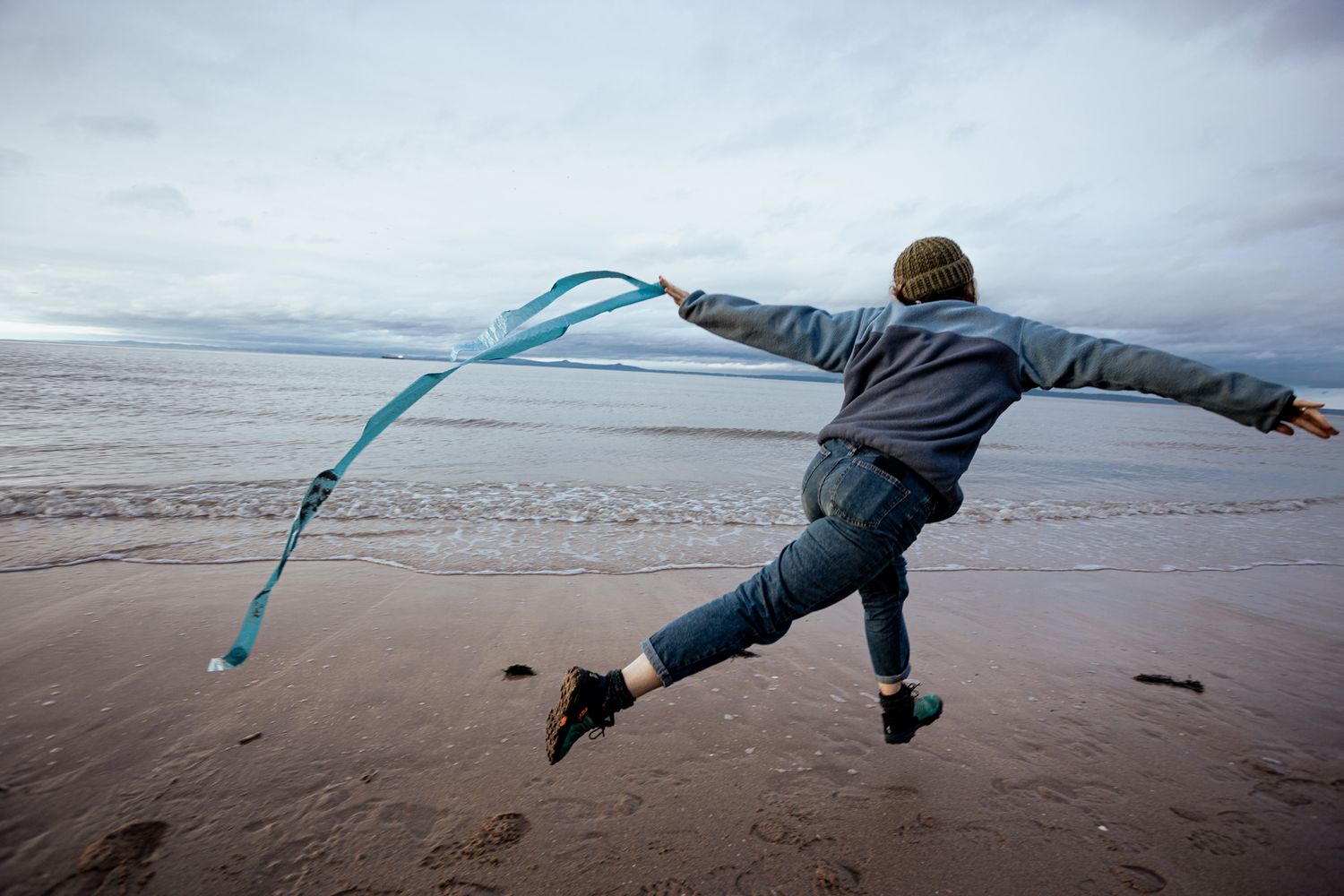 A woman with a blue jumper & jeans is running with a ribbon of blue plastic in front of a blue ocean & grey sky