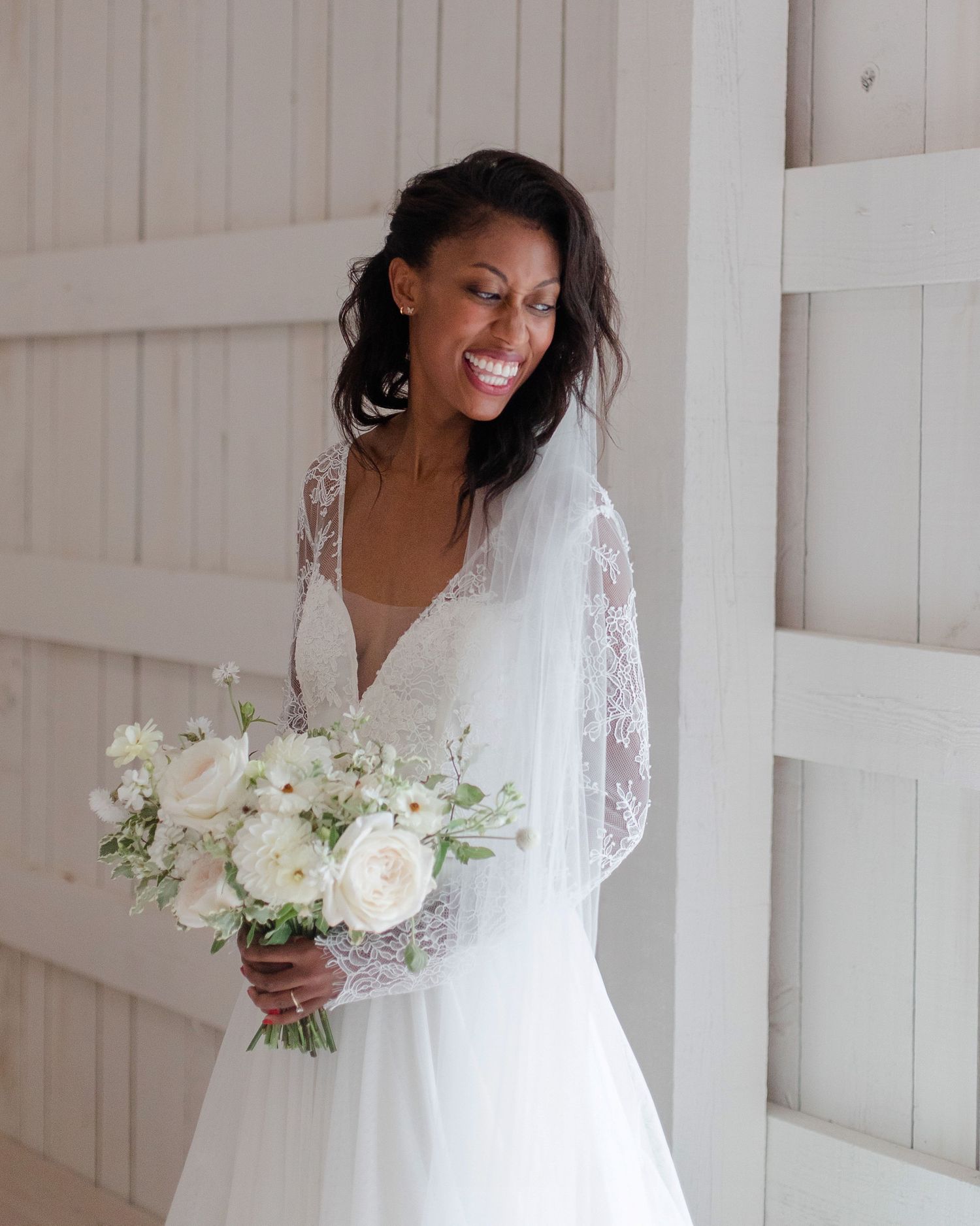 A bride in a white lace gown holds a bouquet of white roses against a white wooden backdrop.
