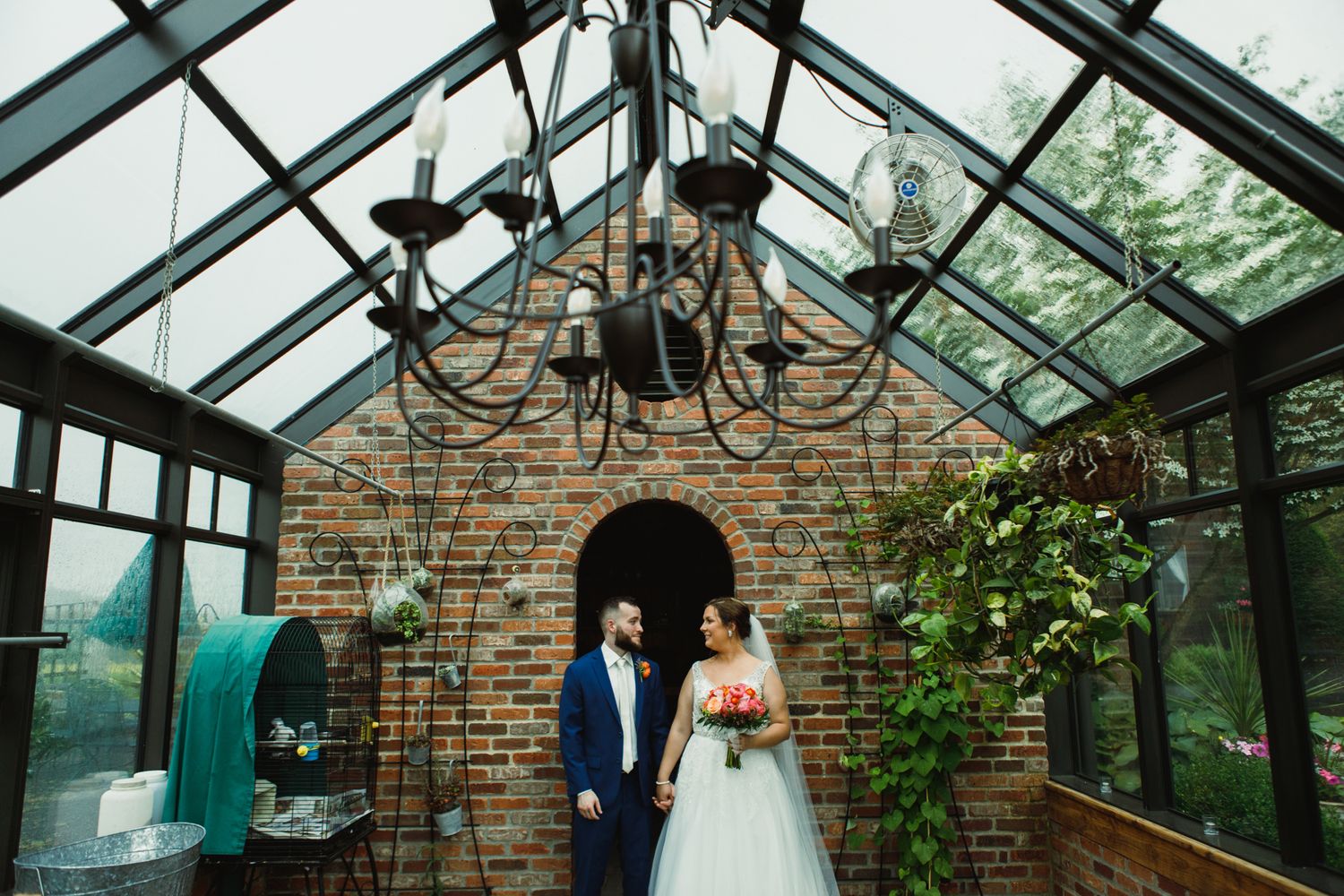 Wedding couple stands under glass ceiling in rustic brick conservatory with chandelier.