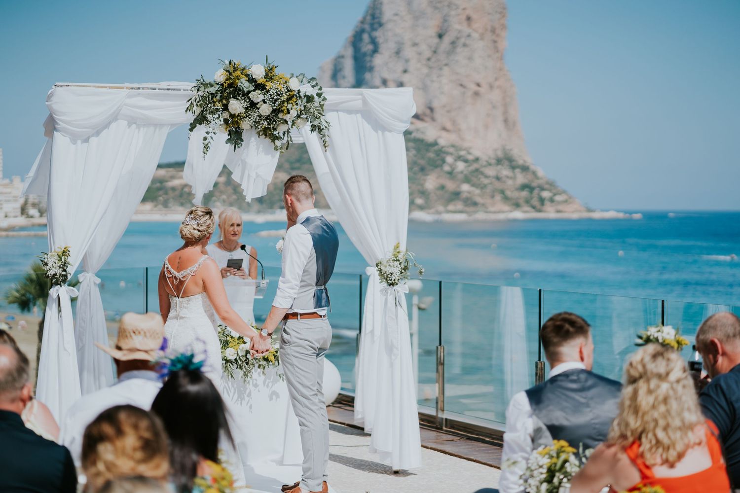 Beach wedding ceremony setup with white arch and chairs overlooking dramatic coastal rock formation.
