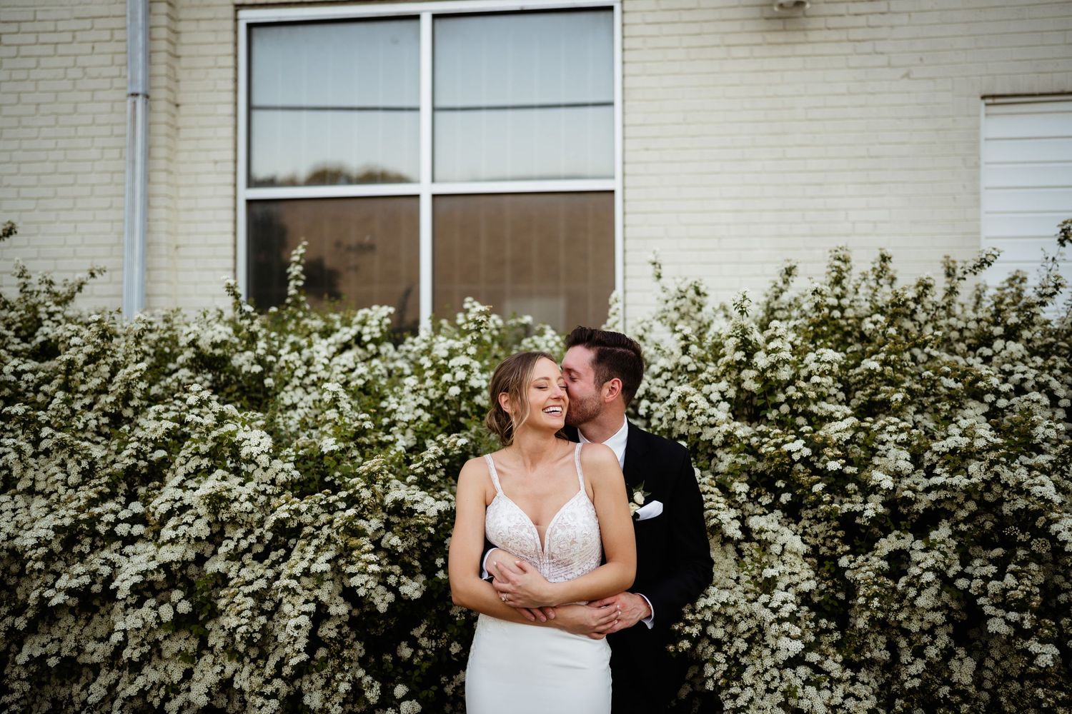 Wedding couple embraces against white brick wall and greenery.