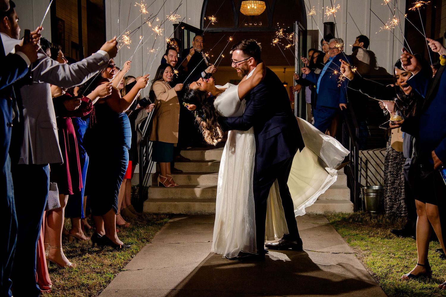 Couple shares romantic moment while guests hold sparklers during nighttime wedding exit.