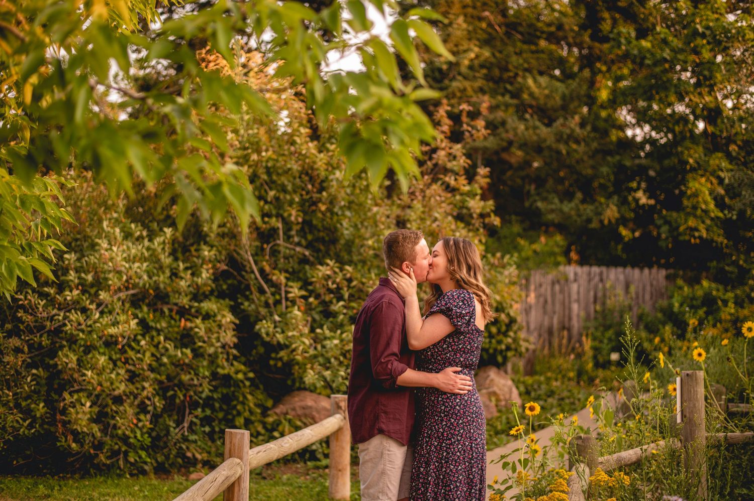 Couple embraces near wooden fence with lush greenery backdrop.