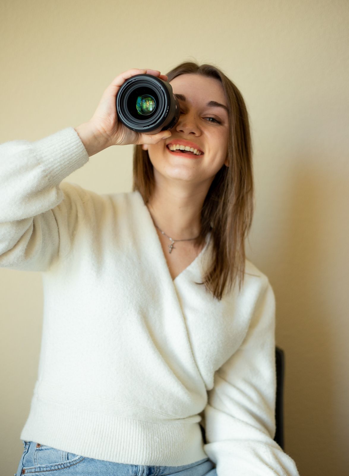 Creative self-portrait holding camera lens in white sweater against beige background.