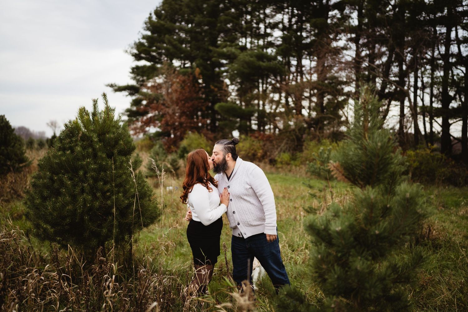 A romantic couple embraces among evergreen trees on a cloudy day.