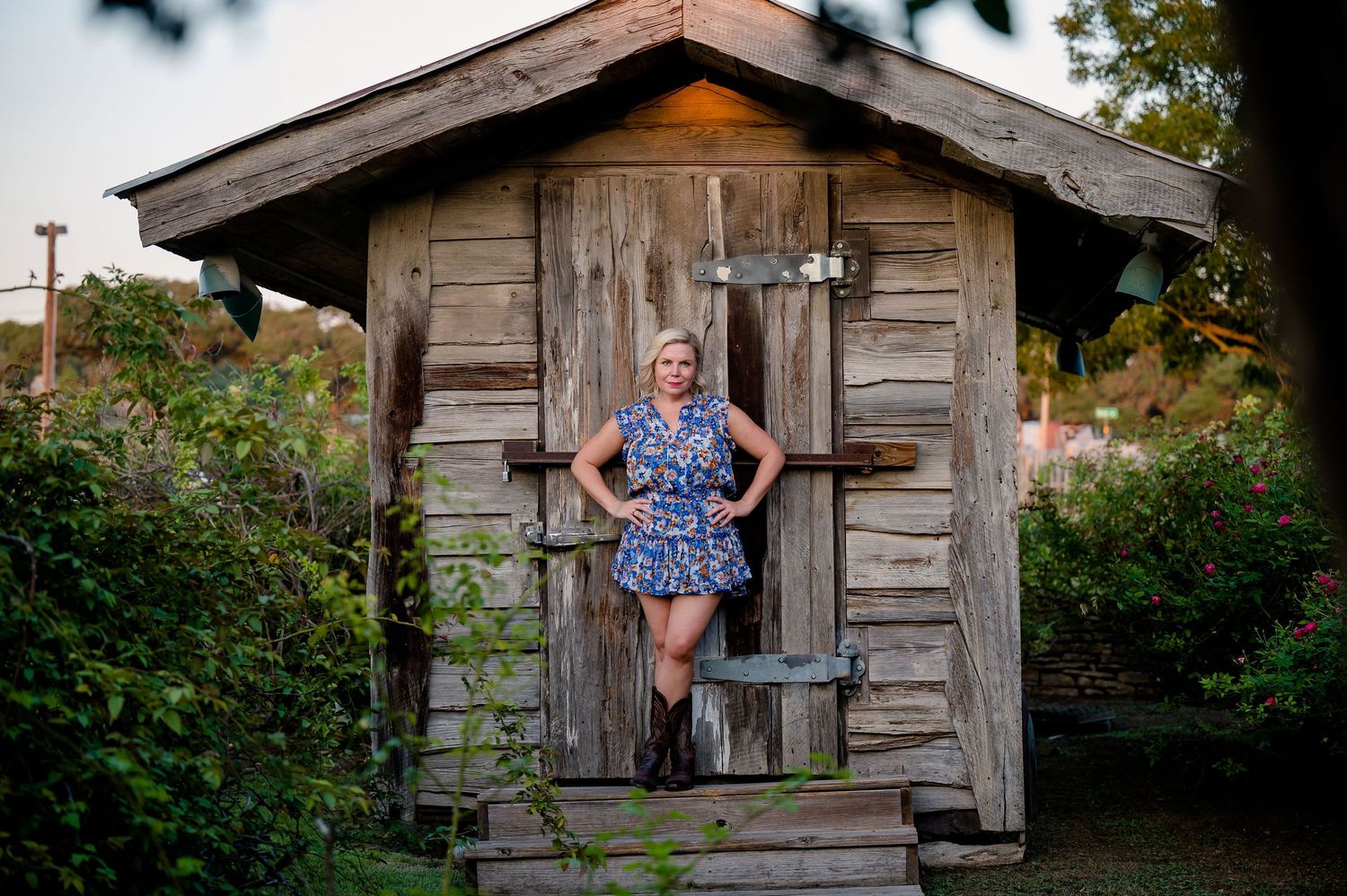 A woman stands in the doorway of a weathered wooden garden shed surrounded by greenery.