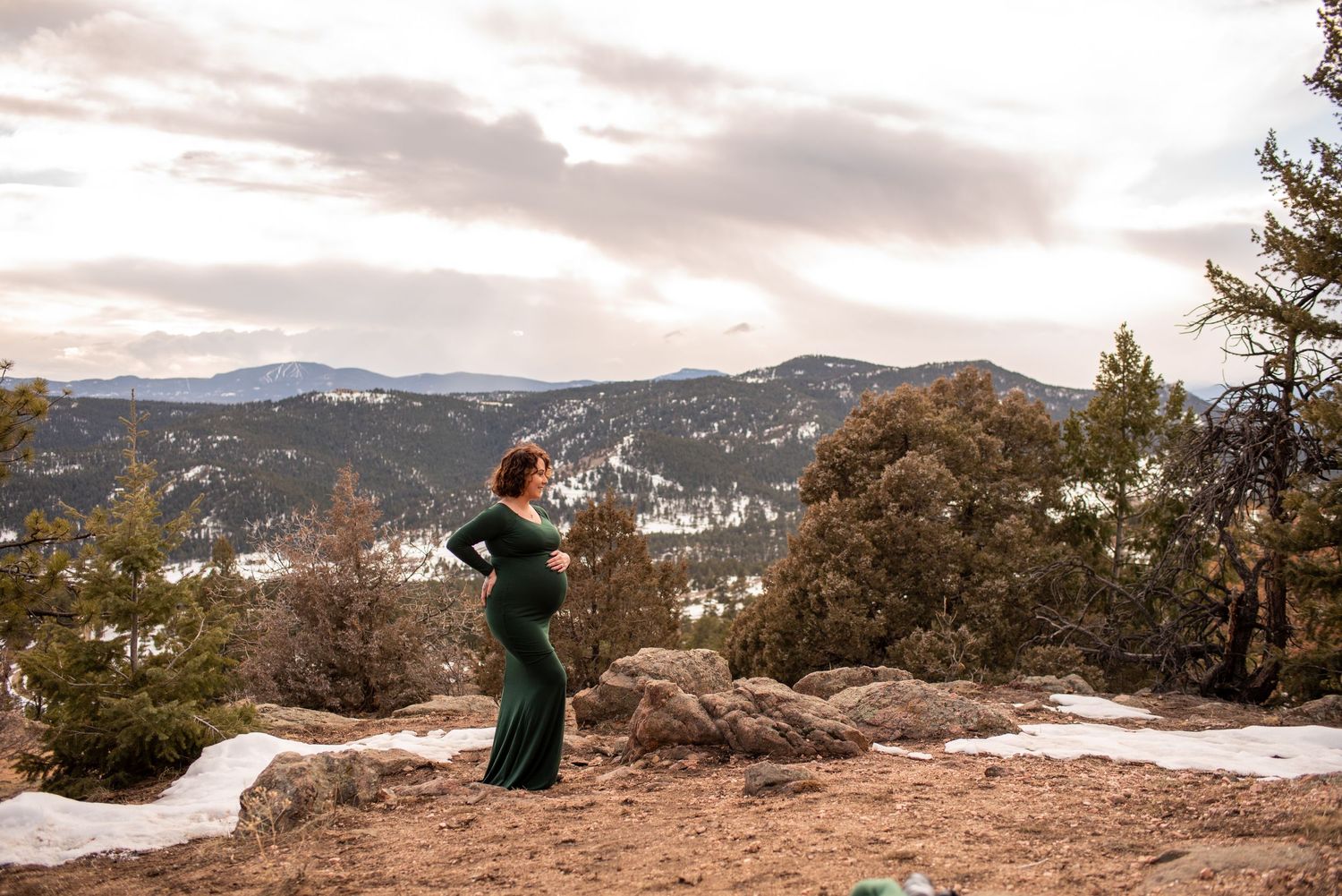 Person in green dress standing on snowy mountain overlook with dramatic cloudy sky.