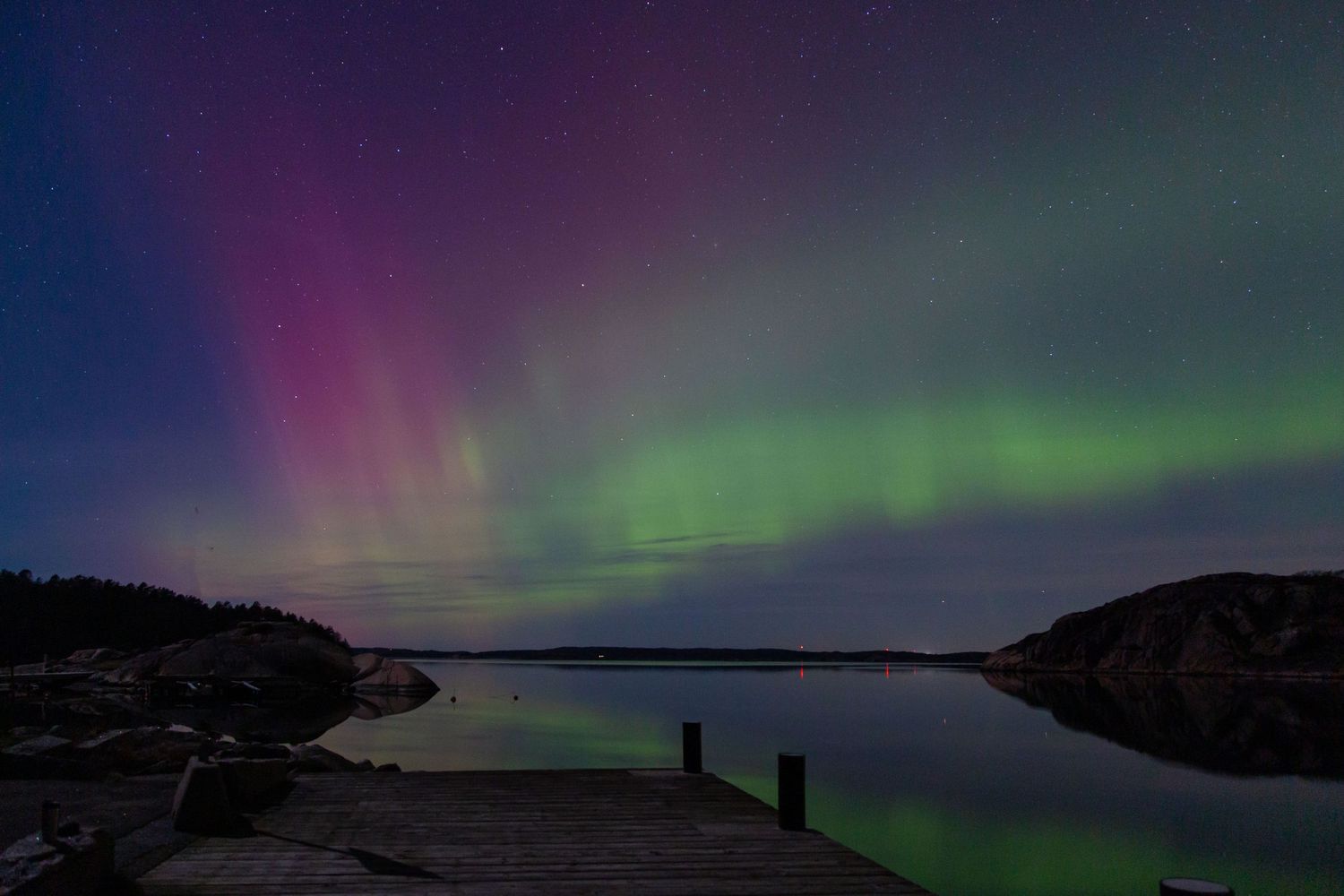 Colorful aurora borealis lights dance over calm lake with dock silhouette.