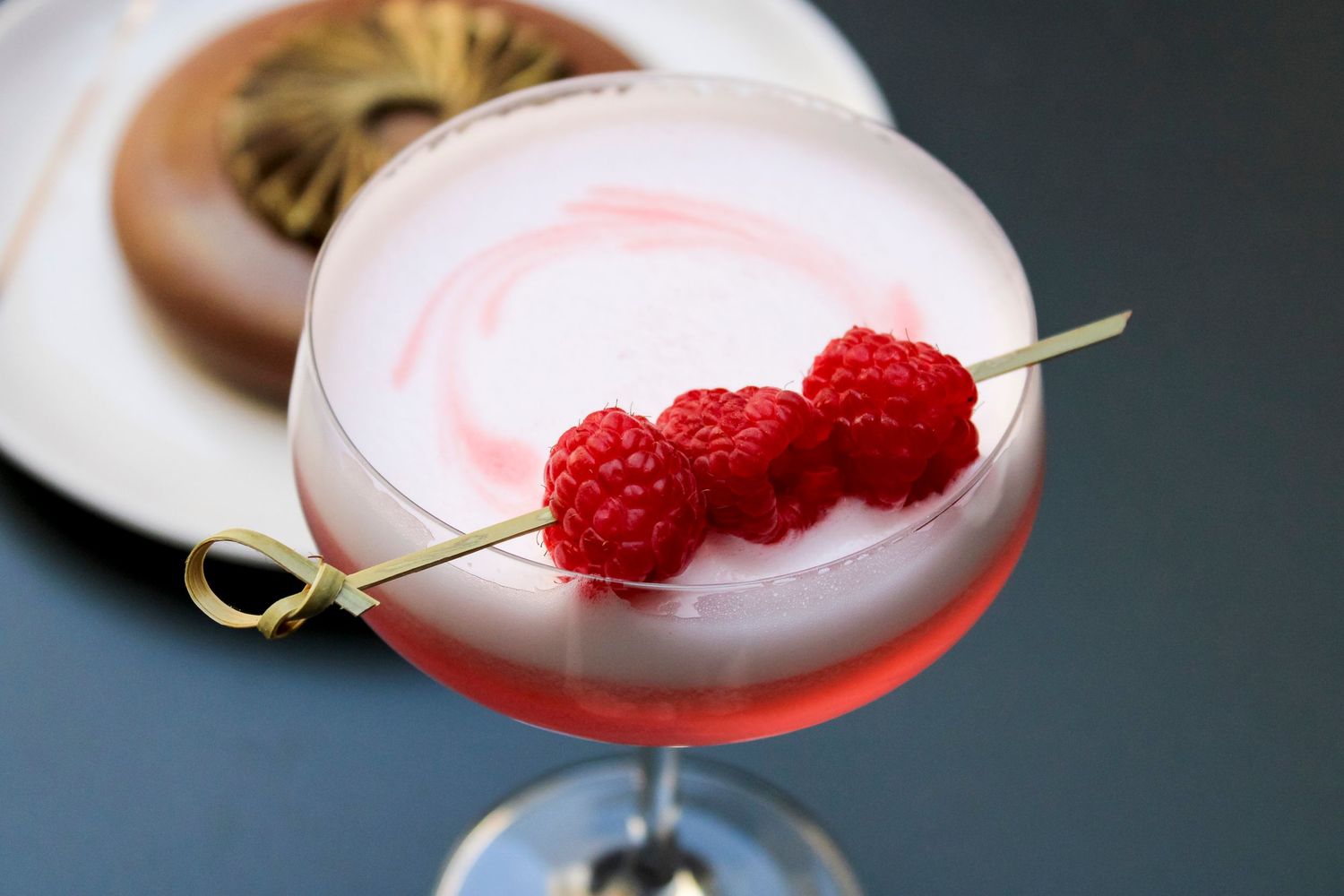 Pink cocktail garnished with fresh raspberries and citrus twist in a coupe glass.