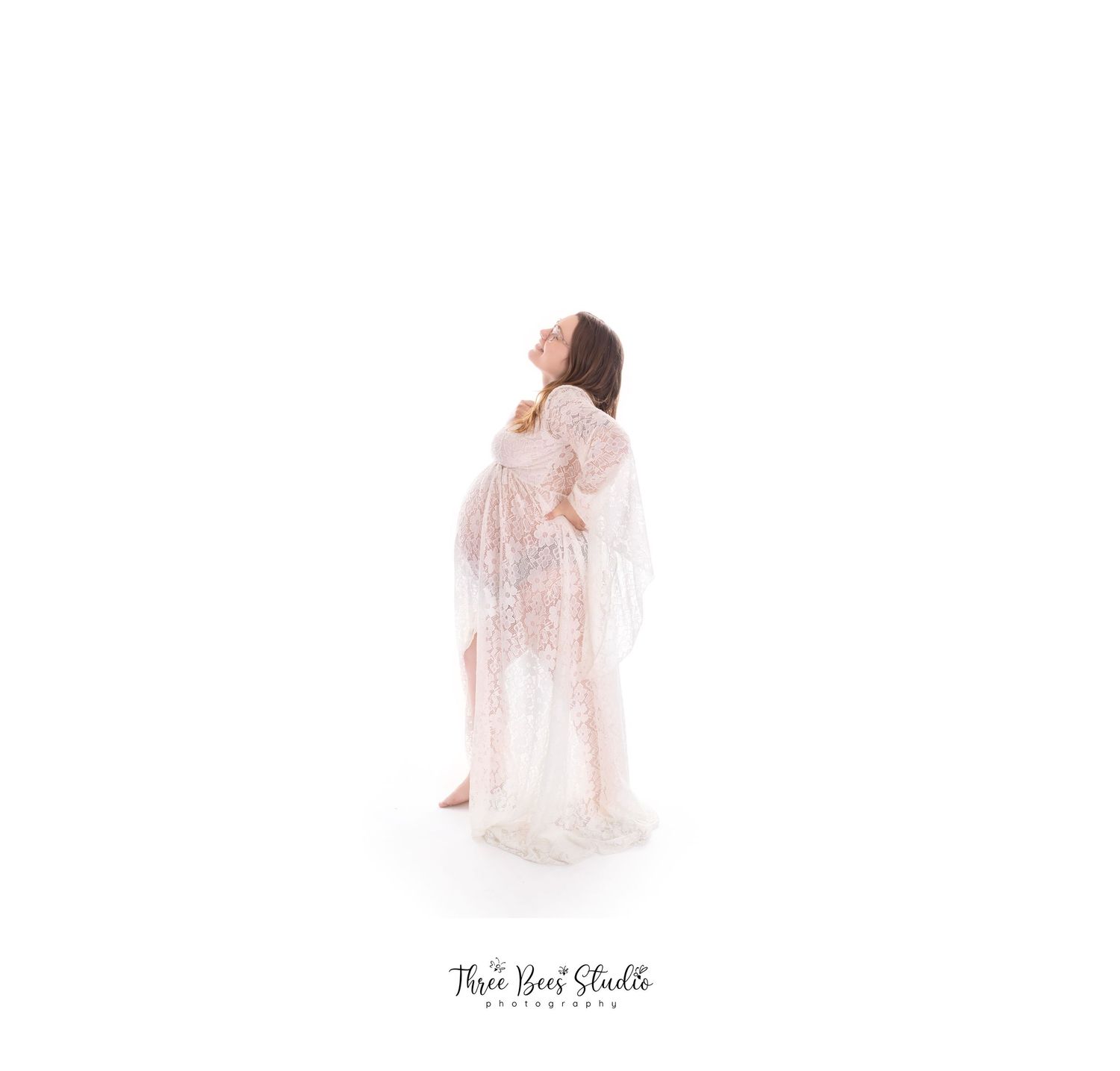 Ethereal maternity portrait with woman in flowing white lace gown against white background.