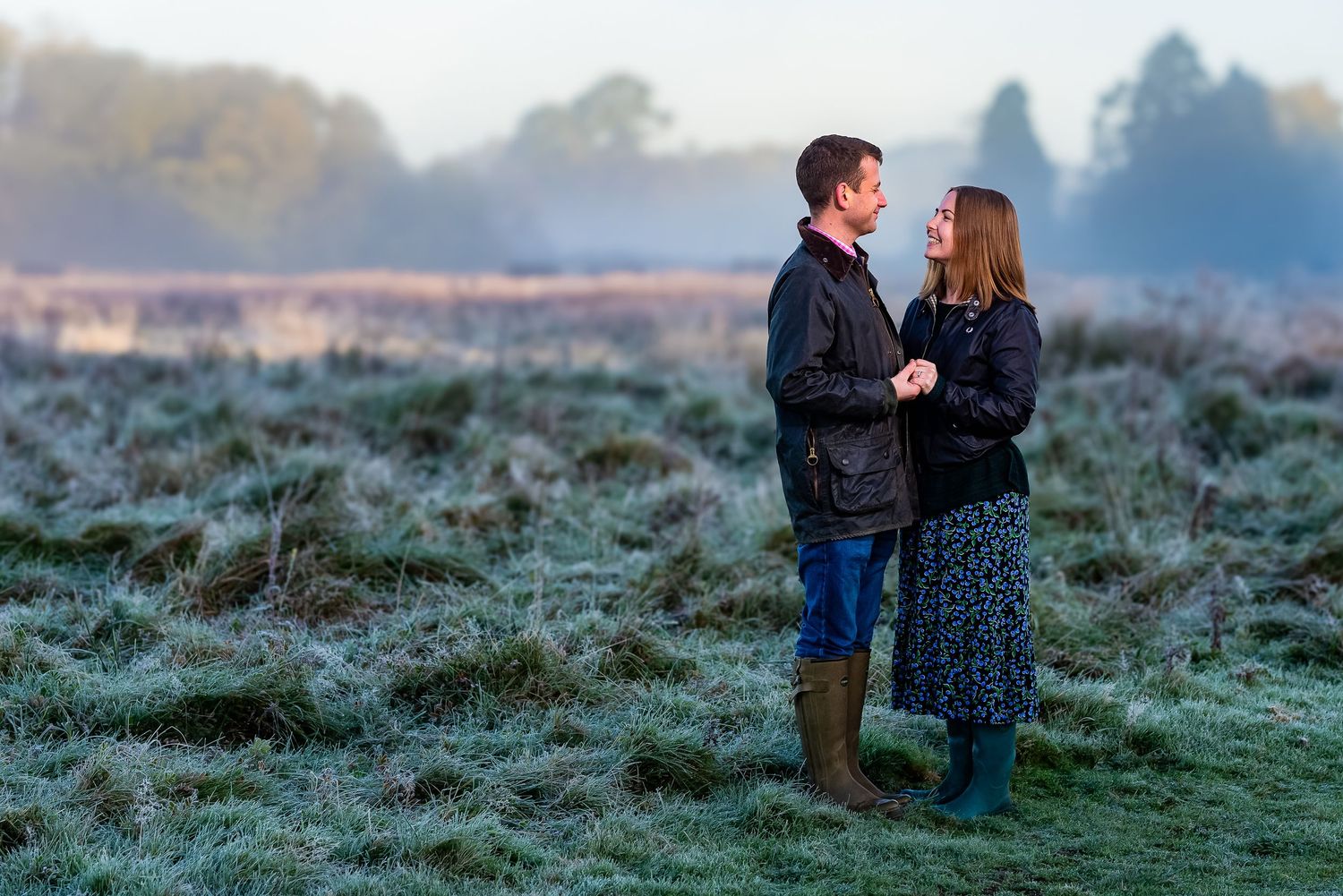 A couple stands close together on a misty field during golden hour.