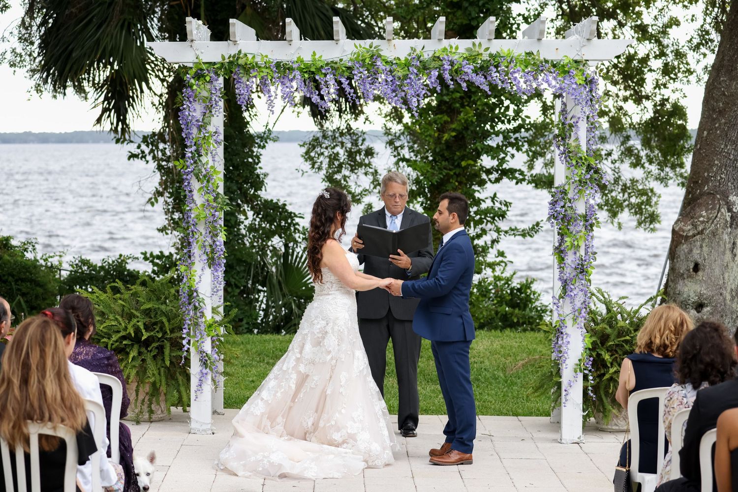 Wedding ceremony under white pergola decorated with purple flowers beside waterfront.