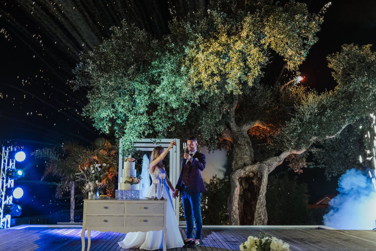 Outdoor wedding reception with colorfully lit trees and elevated DJ booth.