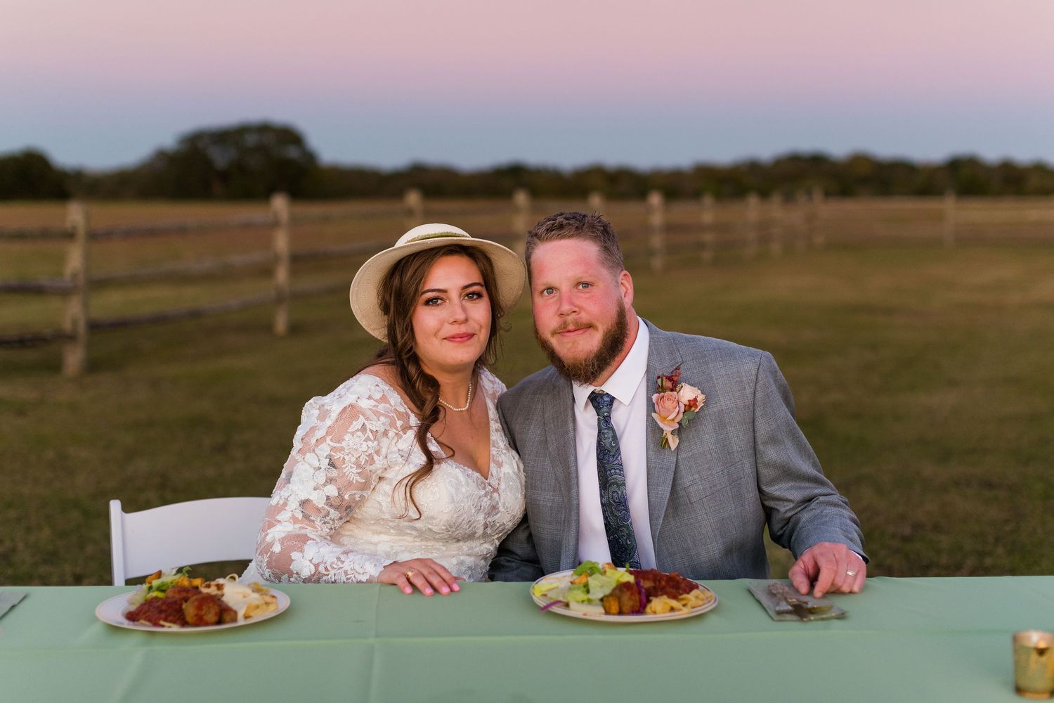 Newlyweds sitting at an outdoor reception table during sunset with dinner plates.