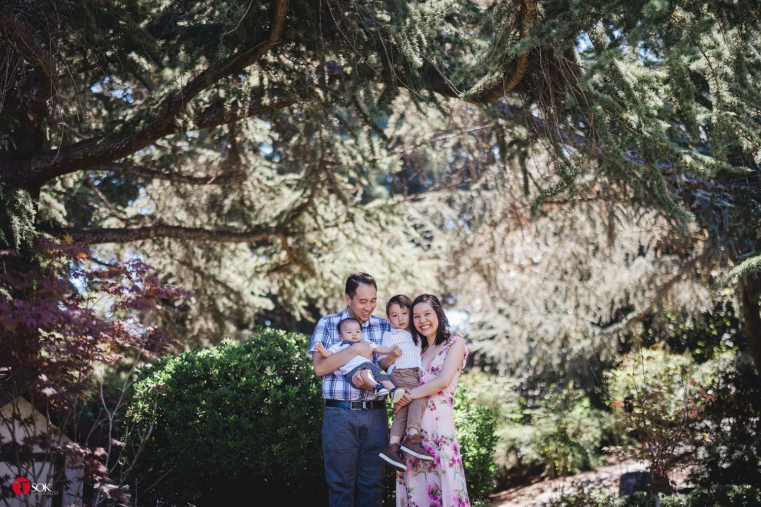 A family poses together under a tree in an outdoor garden setting on a sunny day.