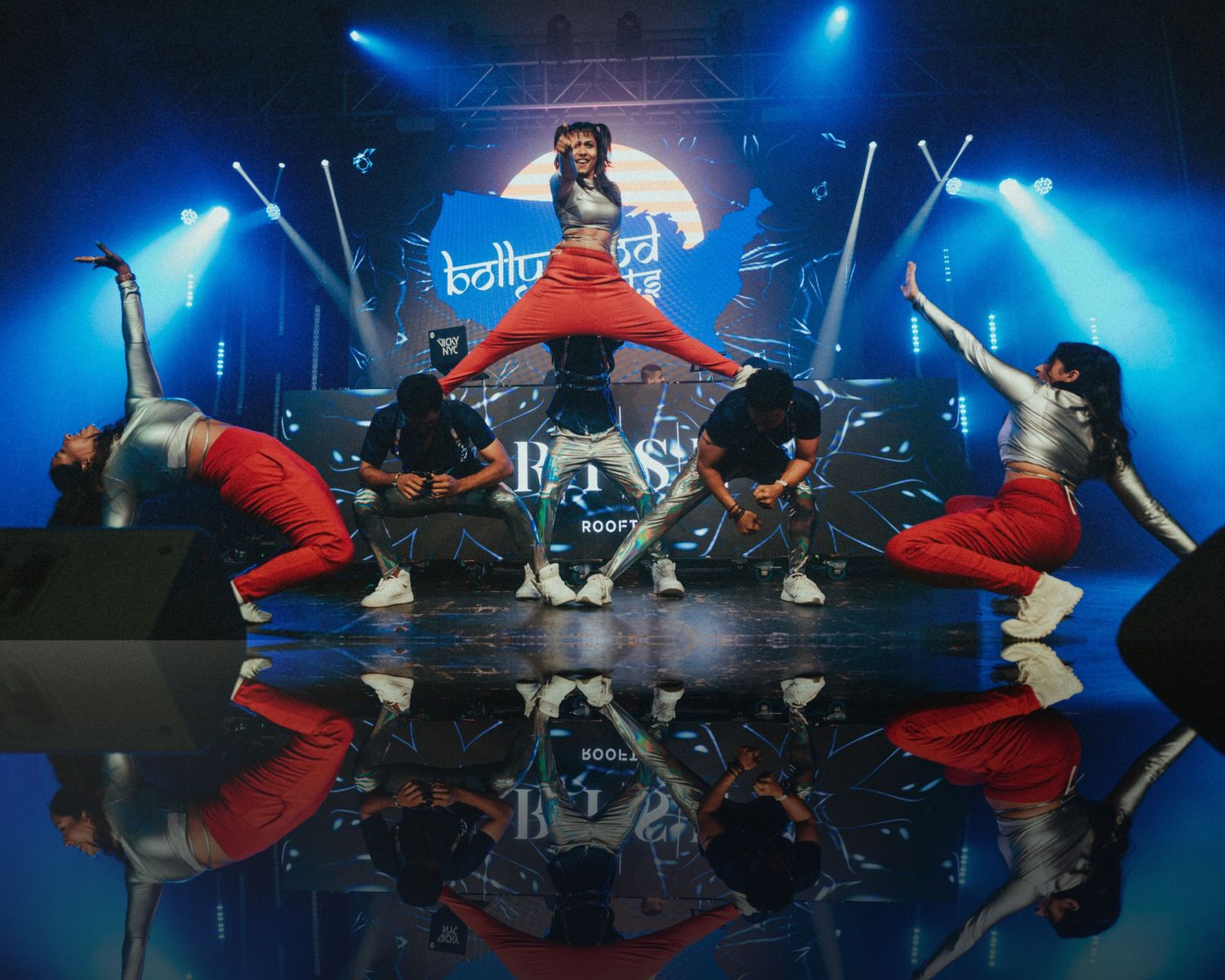 Reflective stage floor mirrors dancers in red pants performing synchronized movements under blue lights.