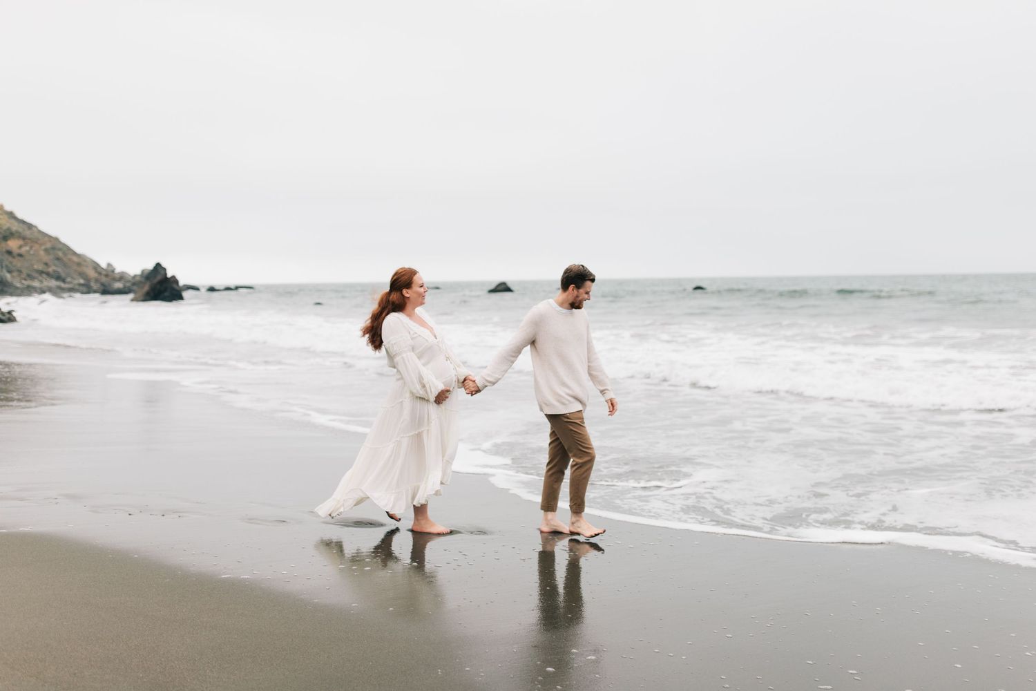 Couple walks hand in hand along misty beach shore in casual wedding attire.
