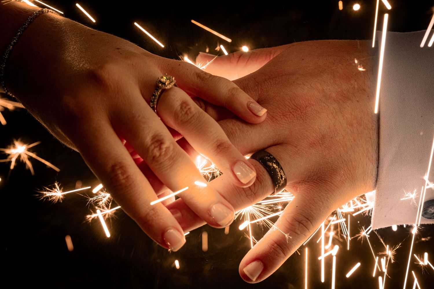 Two hands displaying wedding rings sparkle against a backdrop of glowing sparklers.
