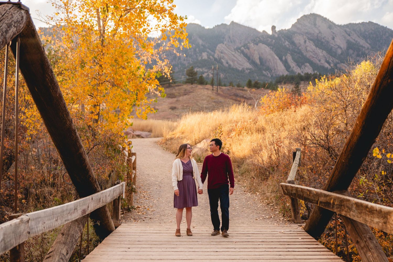 Couple walking hand in hand on wooden bridge with mountain views and autumn foliage.