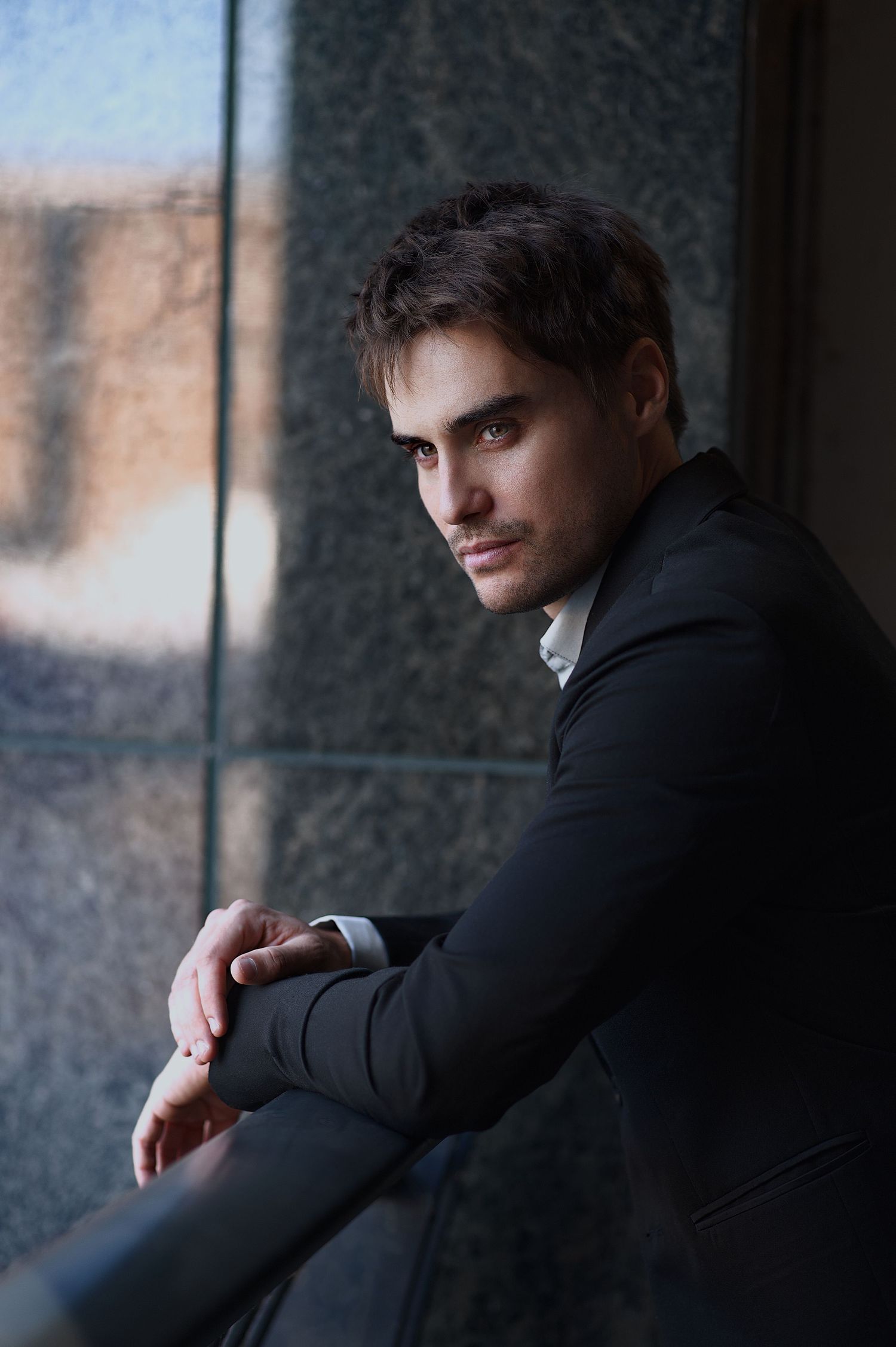A moody portrait of someone in a dark suit looking pensively through a window in dramatic lighting.