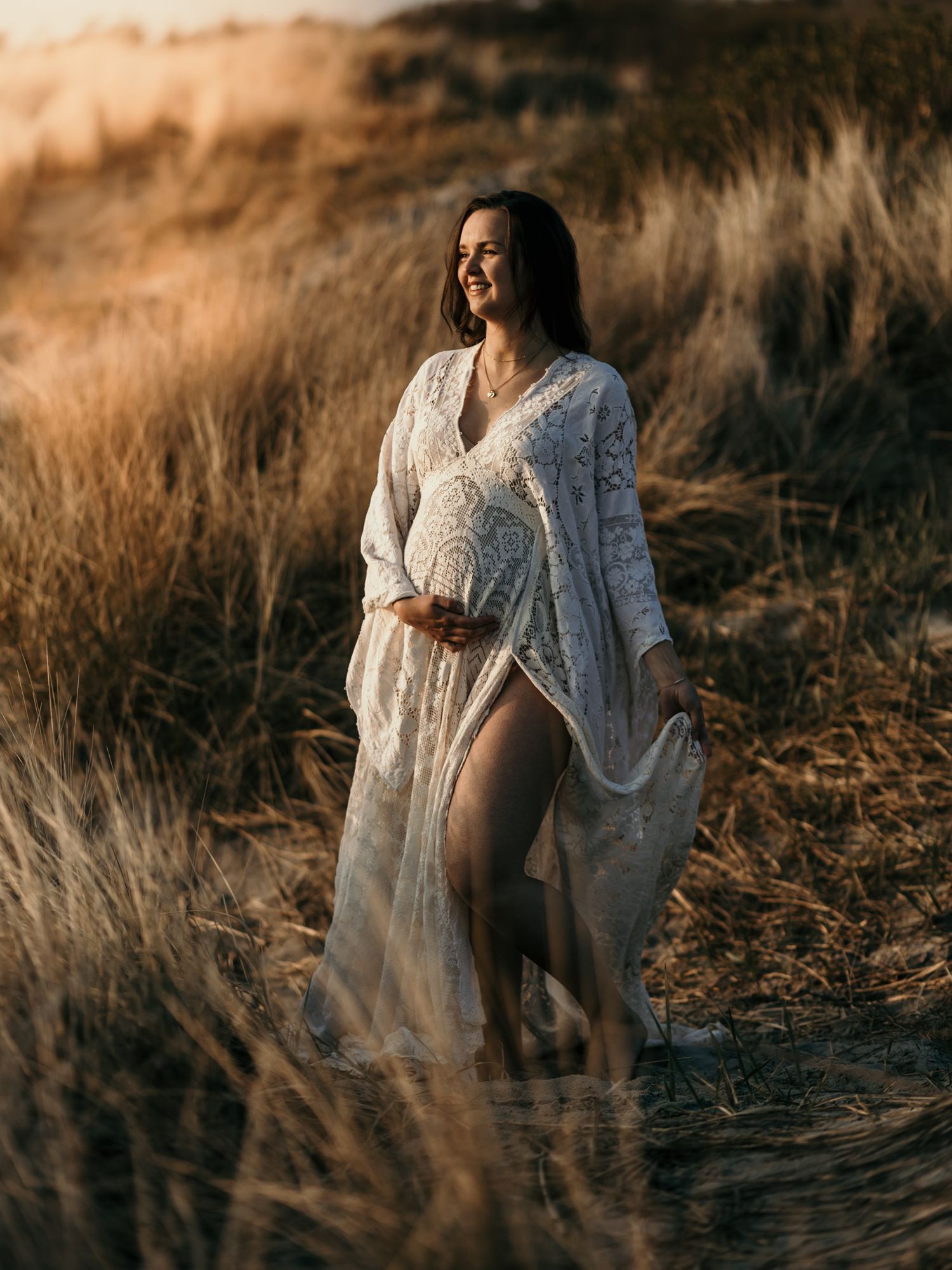 Maternity portrait of woman in flowing white dress standing in golden sunlit field.