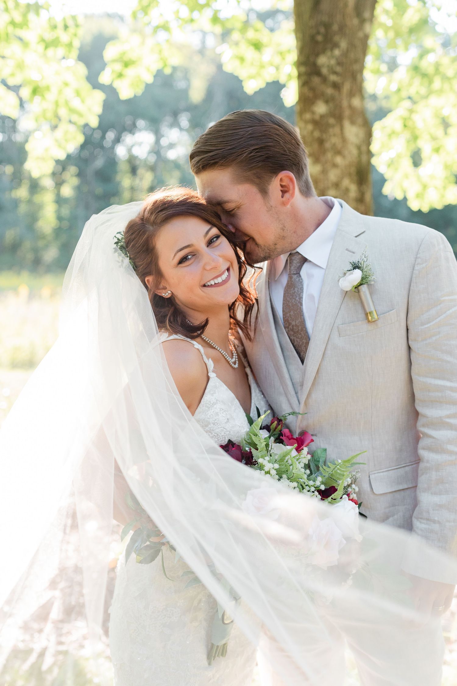 Wedding couple embraces under flowing veil in sunlit garden setting.