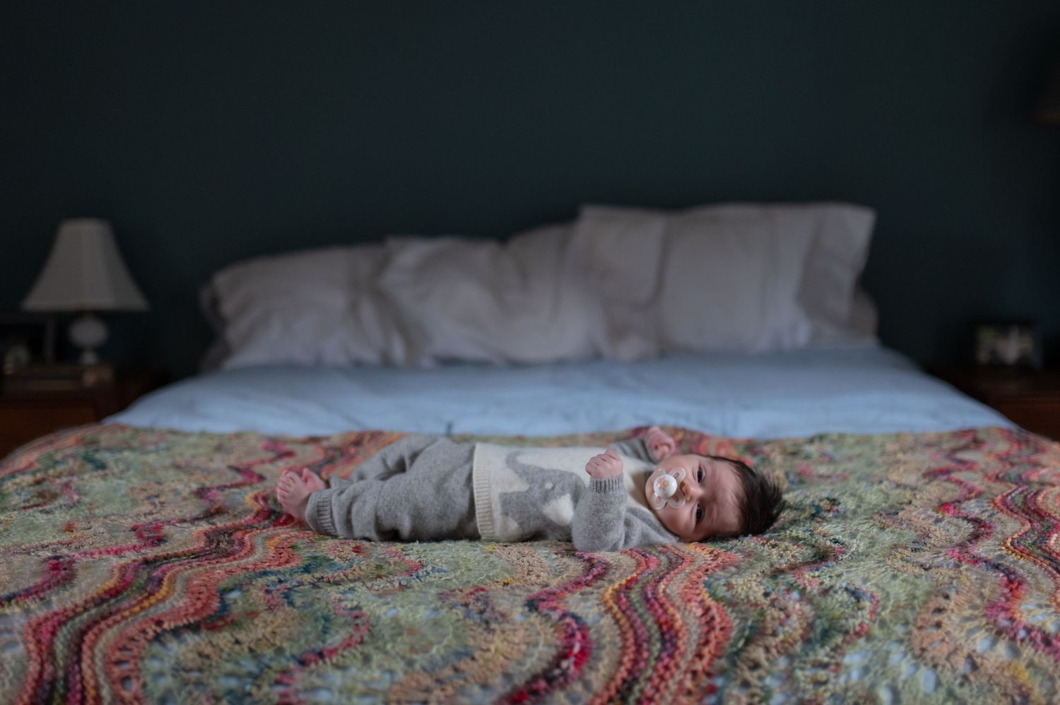 Baby lying peacefully on a colorful patterned bedspread in a dimly lit room.