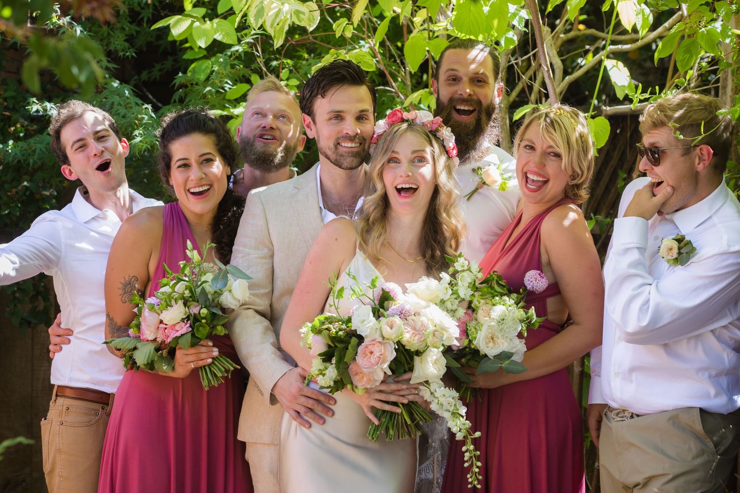 A bridal party in pink and beige attire poses together in a garden setting.