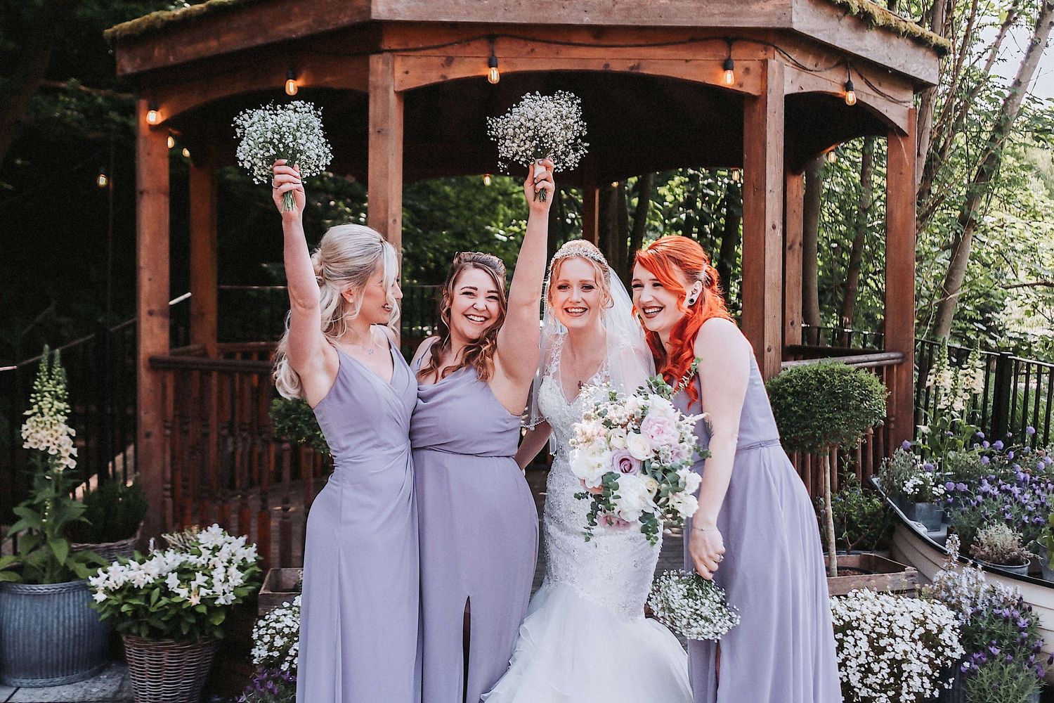 Wedding party in purple dresses poses with bride under rustic gazebo with floral arrangements.