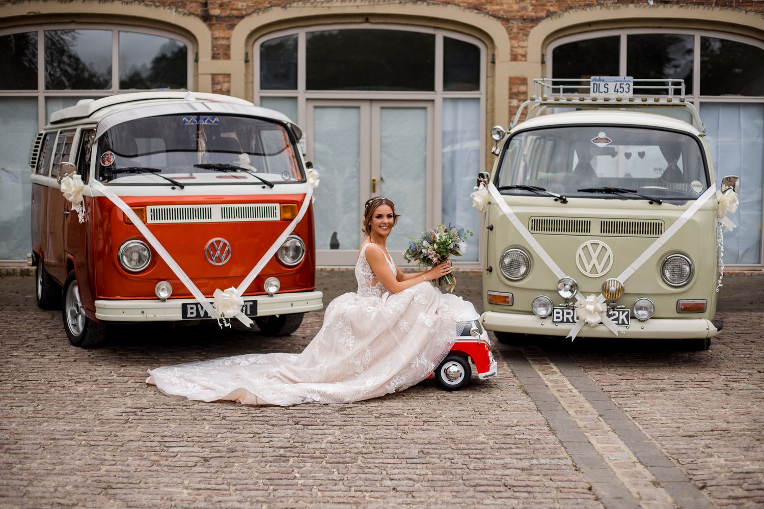Bride in flowing dress poses between classic orange and beige VW campervans.