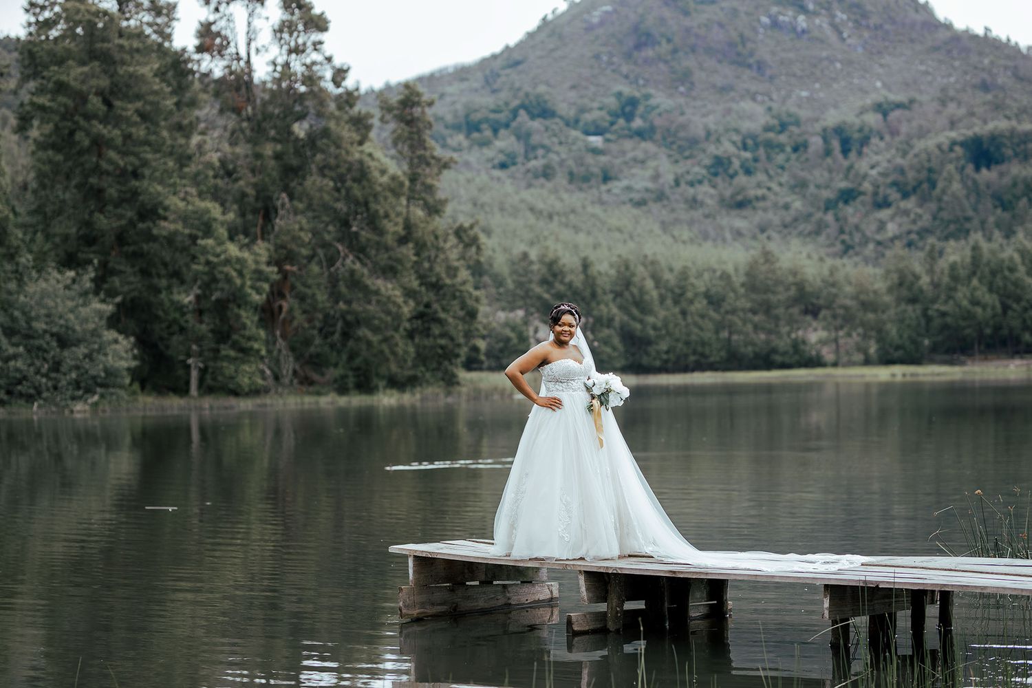 Bride in white wedding gown poses on wooden dock overlooking serene mountain lake.