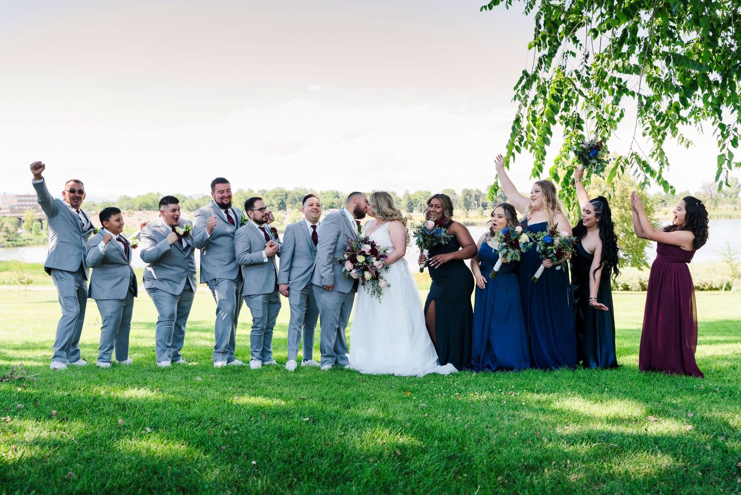 Wedding party in blue and navy attire poses together on grassy hill for group photo.