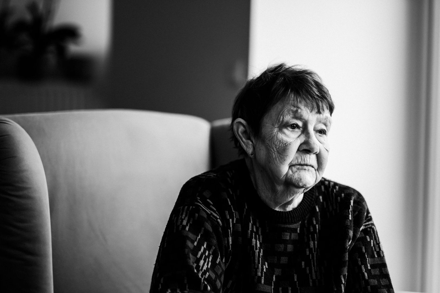 Dramatic black and white portrait of senior woman seated in armchair by window.