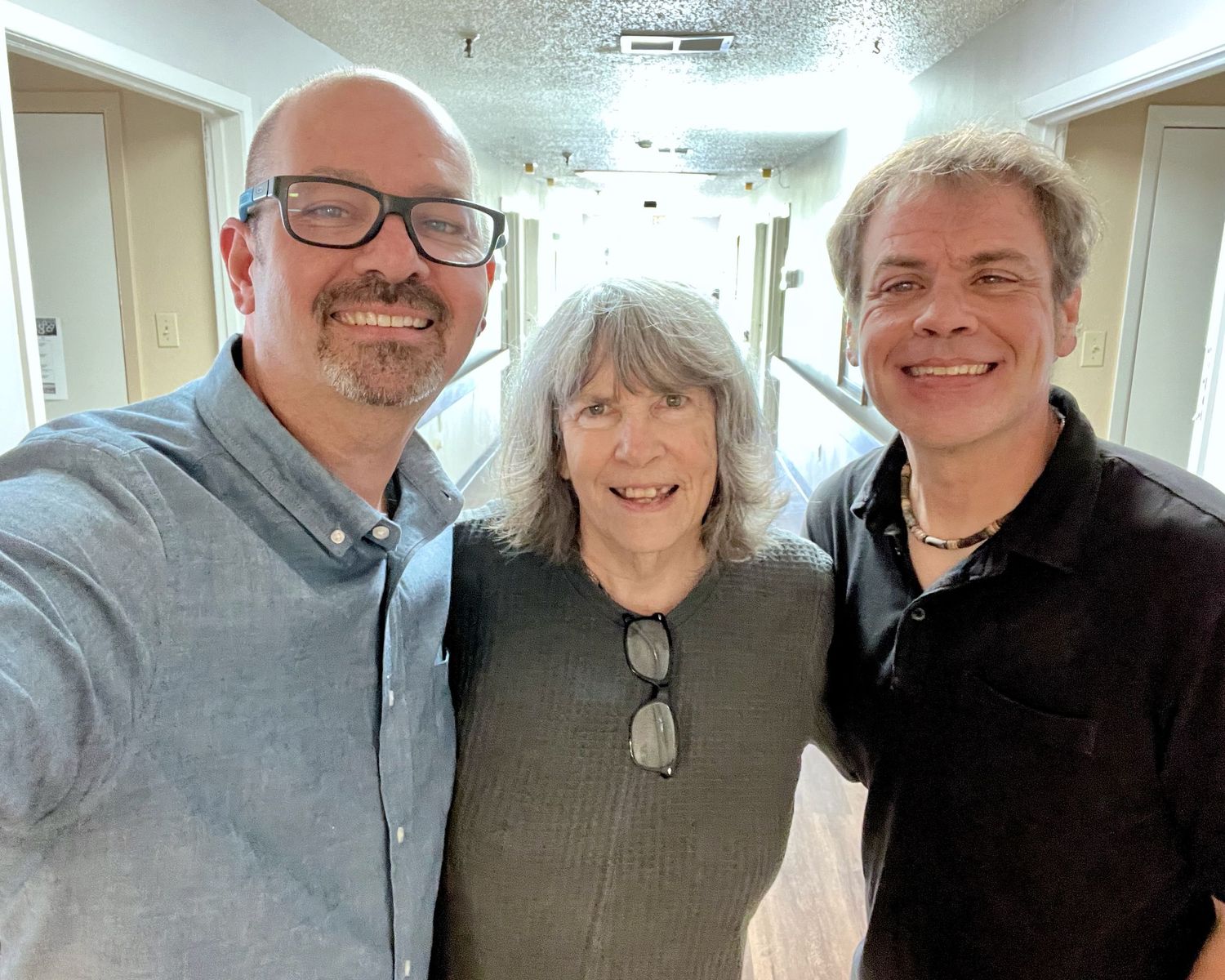 Three smiling people posing for a selfie in a bright hotel corridor hallway.