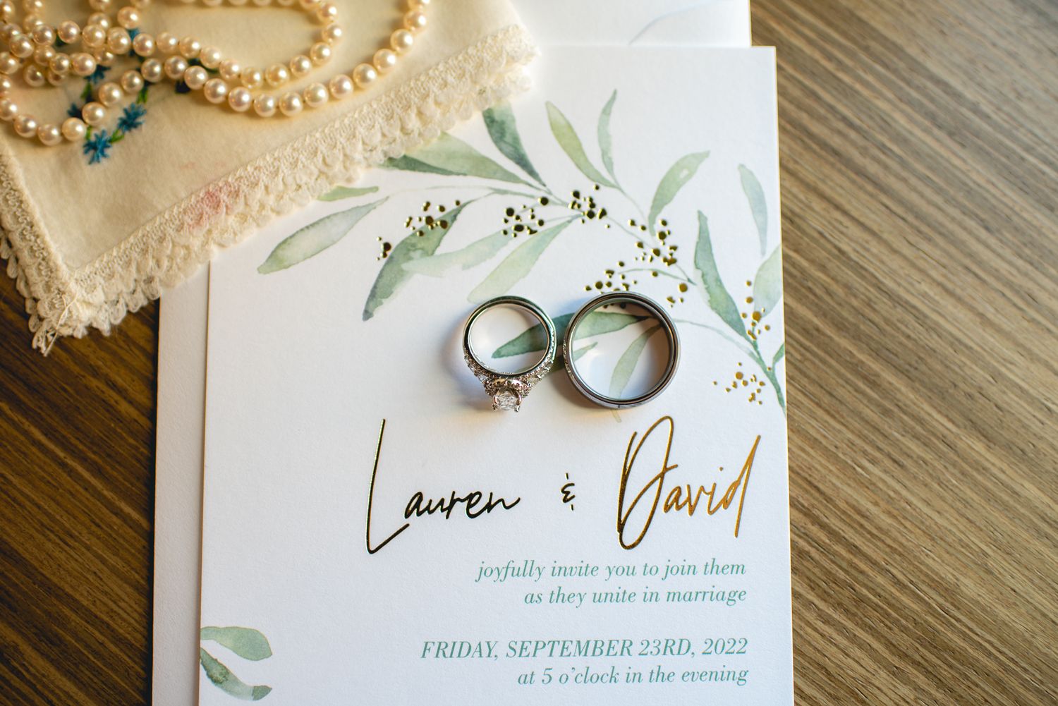 Wedding invitation with silver rings and delicate floral watercolor design on wooden surface.