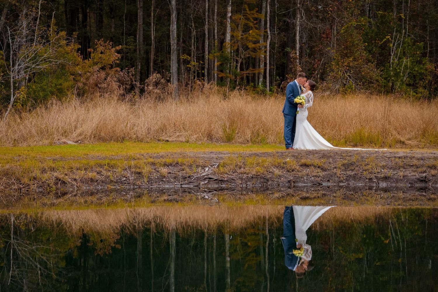 A bride and groom stand by a tranquil lake with their reflection in the water at sunset.