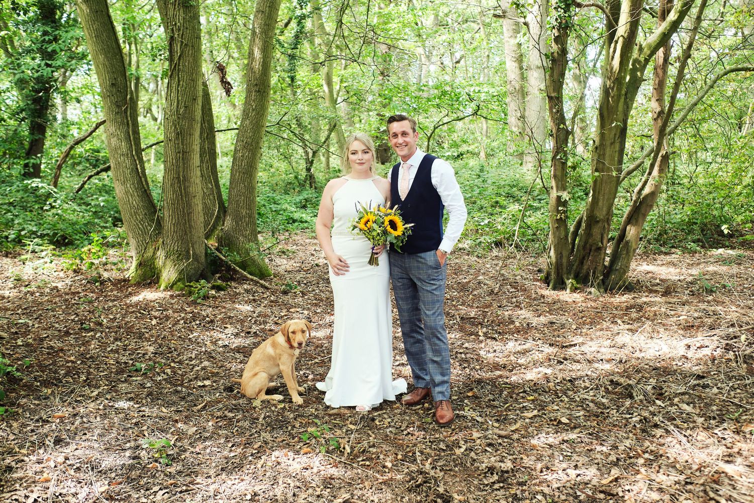 A couple poses for a woodland wedding photo with their dog among trees.