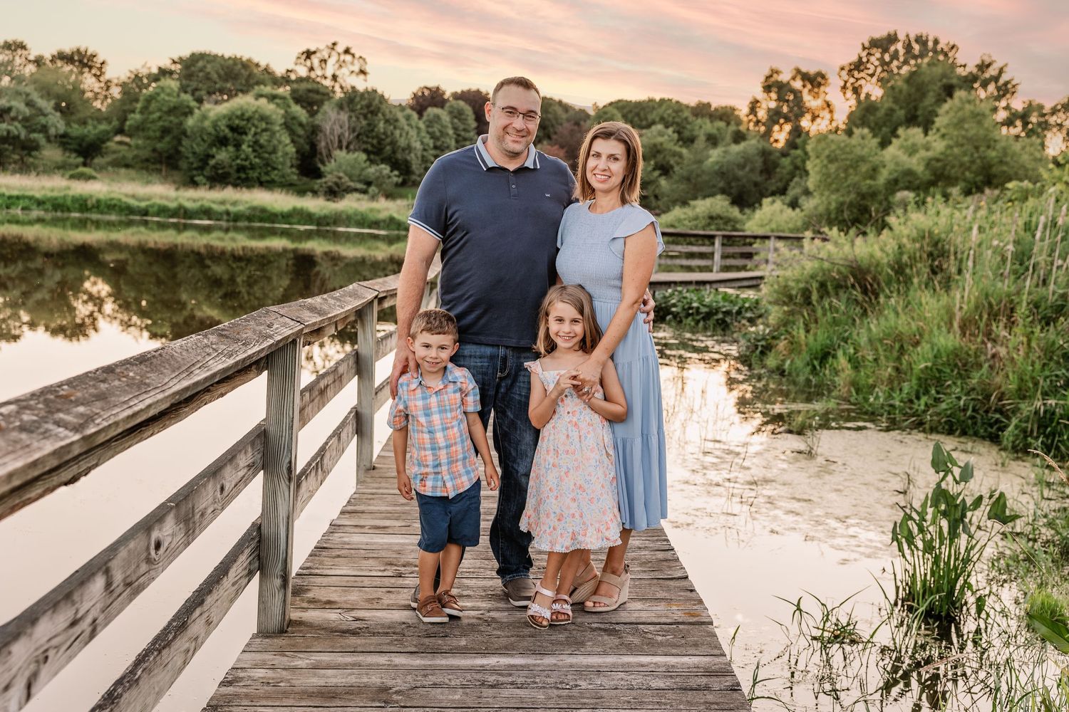Family of four posing together on wooden bridge at sunset over peaceful water.