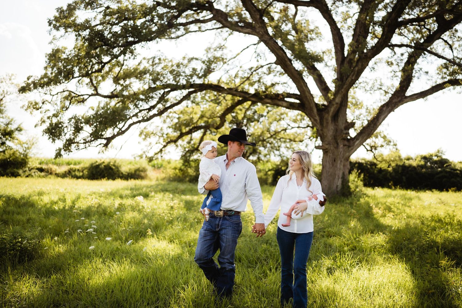 Couple walking hand in hand through a sunlit meadow with majestic oak tree in background.