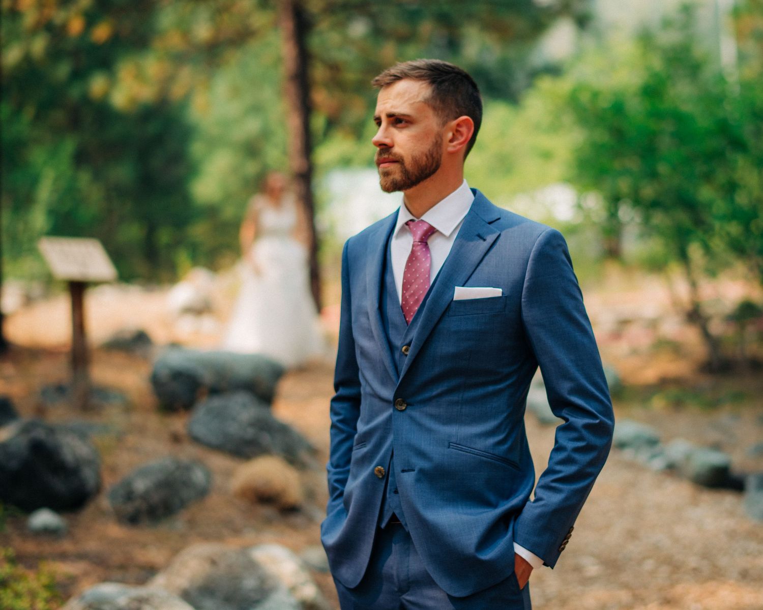 A formal portrait of a person in a fitted navy suit with red tie against a natural outdoor backdrop.
