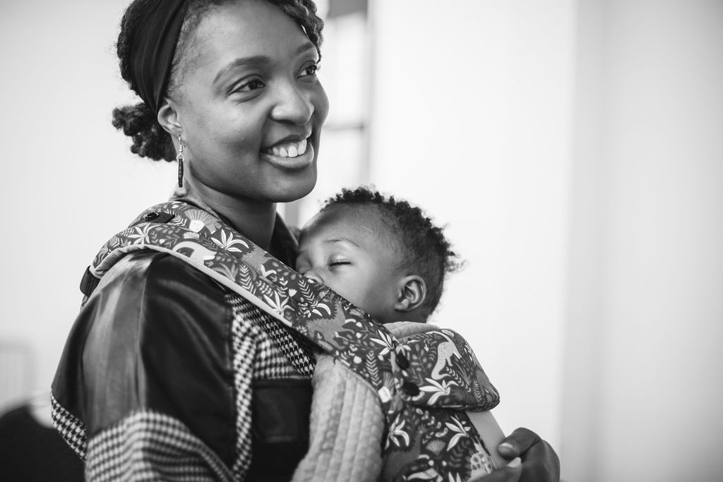 Black and white portrait of a smiling woman wearing a patterned wrap carrying a sleeping baby.