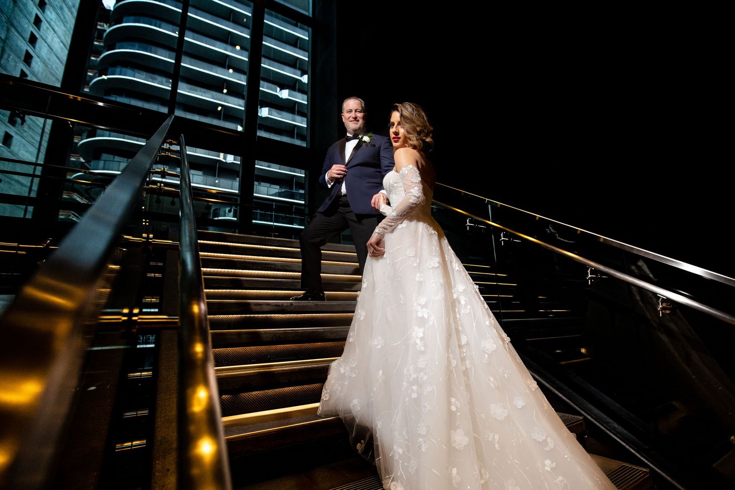 Elegant couple walking down grand staircase at night with dramatic lighting.