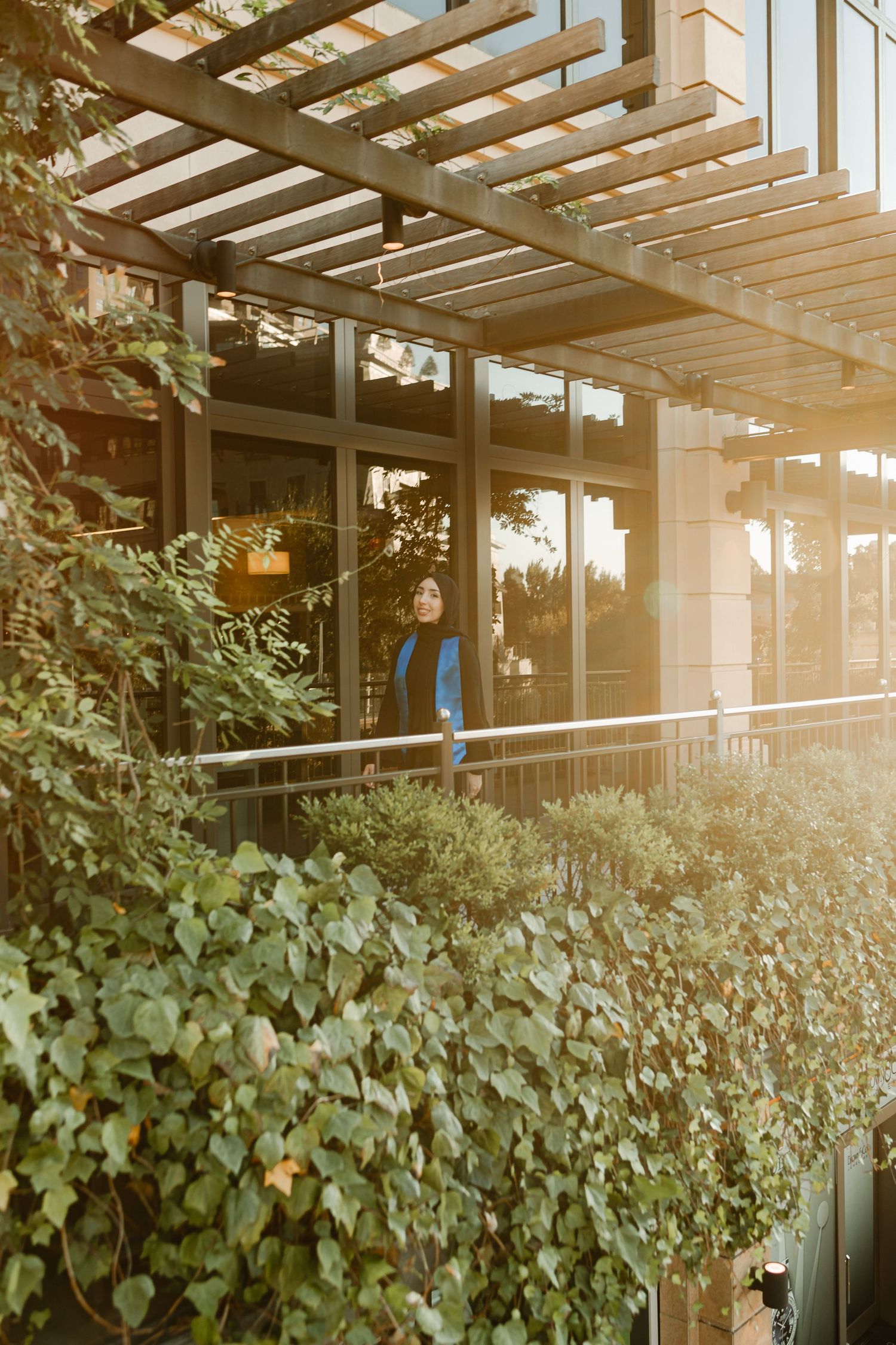 Sunlight streams through a wooden pergola over a modern building entrance with lush greenery.