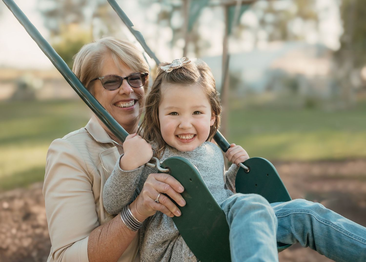 Grandma pushes granddaughter on a swing, both smiling at the camera