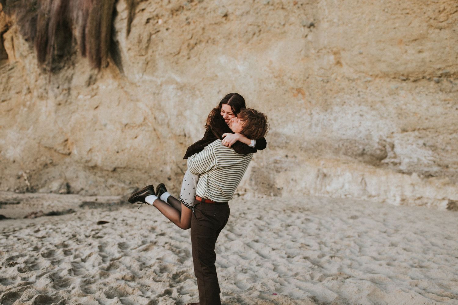 A joyful embrace on a sandy beach with cliffs in the background.